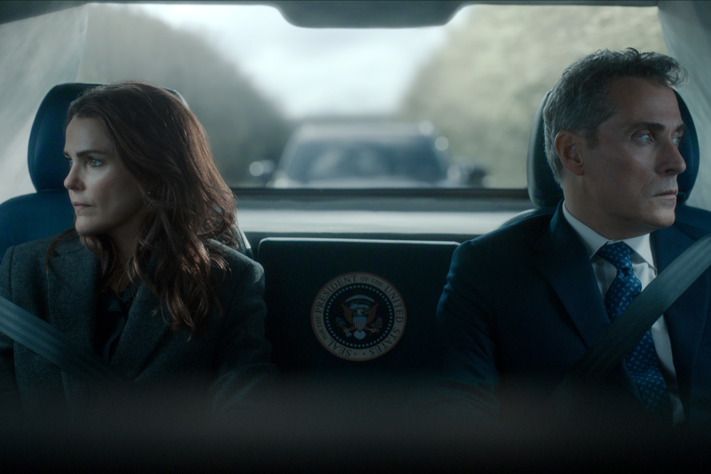 "Two serious people in formal attire sit in the back of a car with a presidential seal, looking out the windows; the environment is tense, with another vehicle visible behind them through the rear window."